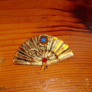 Puede incluir: Un broche de forma de abanico de color dorado con un adorno de pedrer&iacute;a azul y roja.