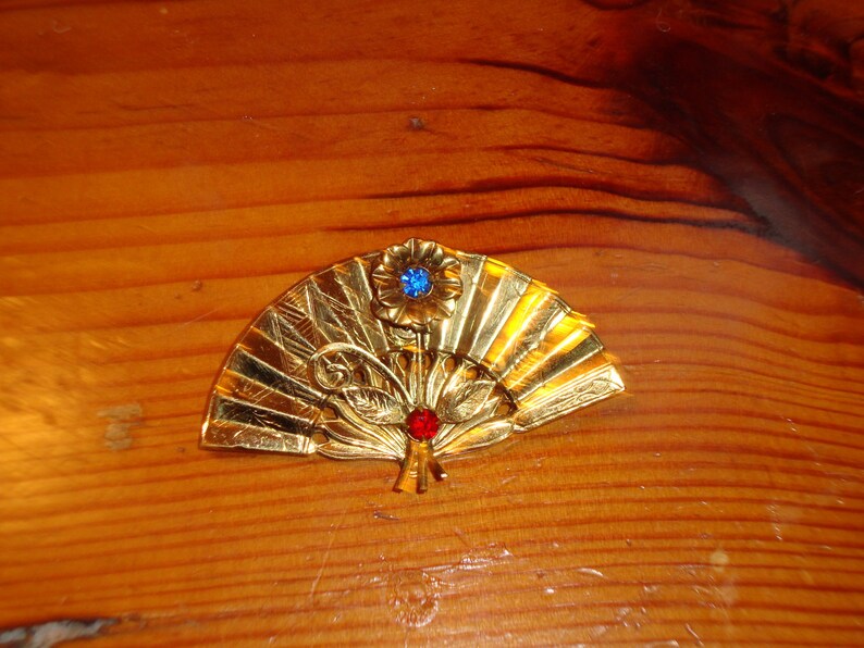 Puede incluir: Un broche de forma de abanico de color dorado con un adorno de pedrer&iacute;a azul y roja.