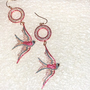 Puede incluir: Un par de pendientes colgantes con un dise&ntilde;o de golondrina rosa y azul con detalles de cristal transparente y rosa. Los pendientes tienen un dise&ntilde;o circular con cristales transparentes y rosas.