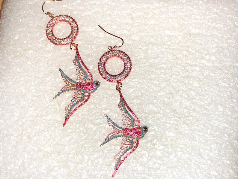 Puede incluir: Un par de pendientes colgantes con un dise&ntilde;o de golondrina rosa y azul con detalles de cristal transparente y rosa. Los pendientes tienen un dise&ntilde;o circular con cristales transparentes y rosas.