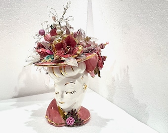 May include: A white ceramic vase shaped like a woman's head with a pink and gold hat decorated with flowers, beads, and crystals.