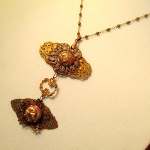 Może przedstawiać: Złoty naszyjnik z filigranowym wisiorem ozdobionym r&oacute;żowym i złotym guzikiem. Wisiorek jest przymocowany do łańcuszka z mniejszym filigranowym wisiorem ozdobionym r&oacute;żowym i złotym guzikiem.