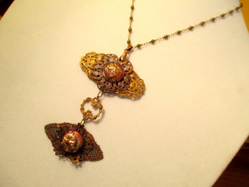 Może przedstawiać: Złoty naszyjnik z filigranowym wisiorem ozdobionym r&oacute;żowym i złotym guzikiem. Wisiorek jest przymocowany do łańcuszka z mniejszym filigranowym wisiorem ozdobionym r&oacute;żowym i złotym guzikiem.