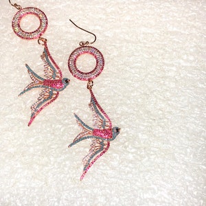 Puede incluir: Un par de pendientes colgantes con un dise&ntilde;o de p&aacute;jaro rosa y azul con detalles de cristal transparente. Los pendientes tienen un dise&ntilde;o circular en la parte superior.