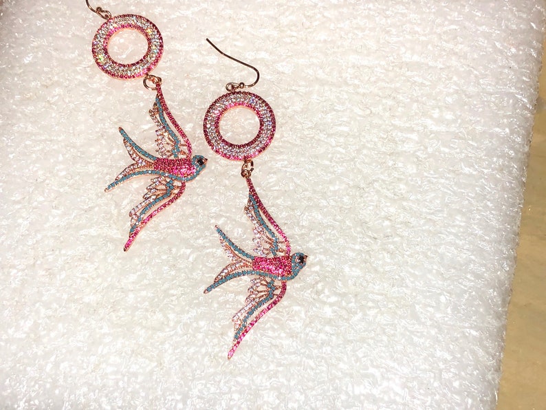 Puede incluir: Un par de pendientes colgantes con un dise&ntilde;o de p&aacute;jaro rosa y azul con detalles de cristal transparente. Los pendientes tienen un dise&ntilde;o circular en la parte superior.