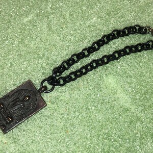 May include: A black chain necklace with a rectangular pendant featuring a detailed image of a person. The pendant is made of metal with a bronze finish. The chain has a magnetic clasp.