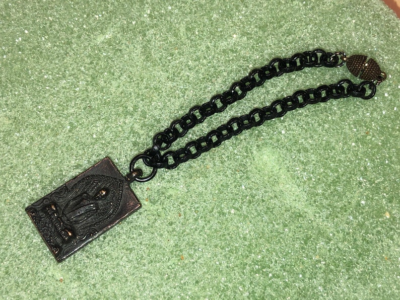 May include: A black chain necklace with a rectangular pendant featuring a detailed image of a person. The pendant is made of metal with a bronze finish. The chain has a magnetic clasp.