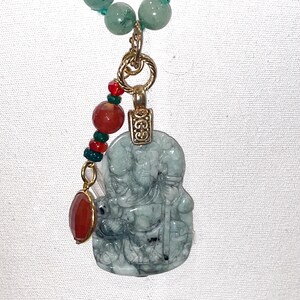 Puede incluir: Un collar con un colgante de jade verde, tallado con la figura de una persona. El colgante est&aacute; unido a una cadena de oro con cuentas rojas y verdes.