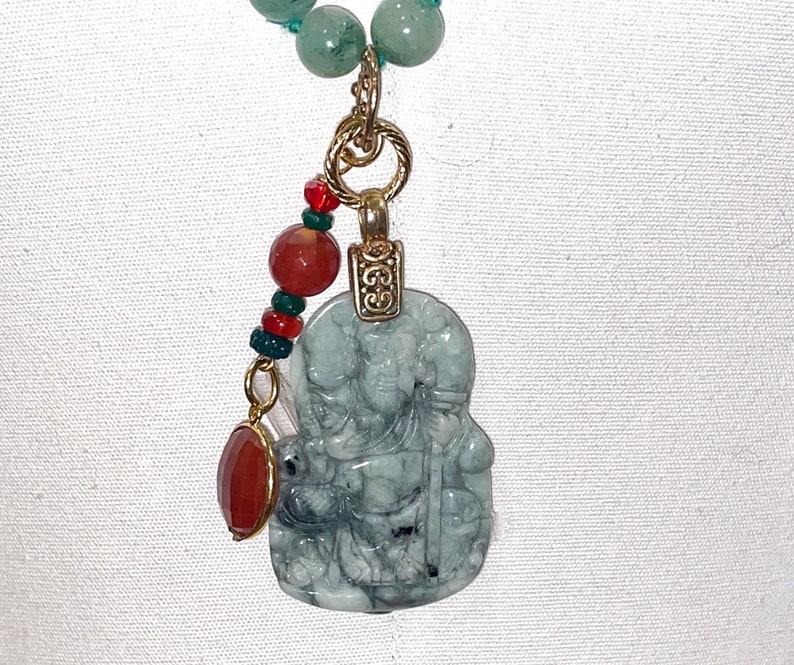 Puede incluir: Un collar con un colgante de jade verde, tallado con la figura de una persona. El colgante est&aacute; unido a una cadena de oro con cuentas rojas y verdes.