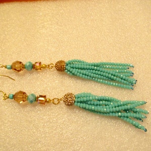 May include: A pair of long, beaded tassel earrings. The earrings feature gold-colored fish hooks, with a series of amber and turquoise beads leading to a cluster of turquoise beads. The tassels are made of small turquoise beads.
