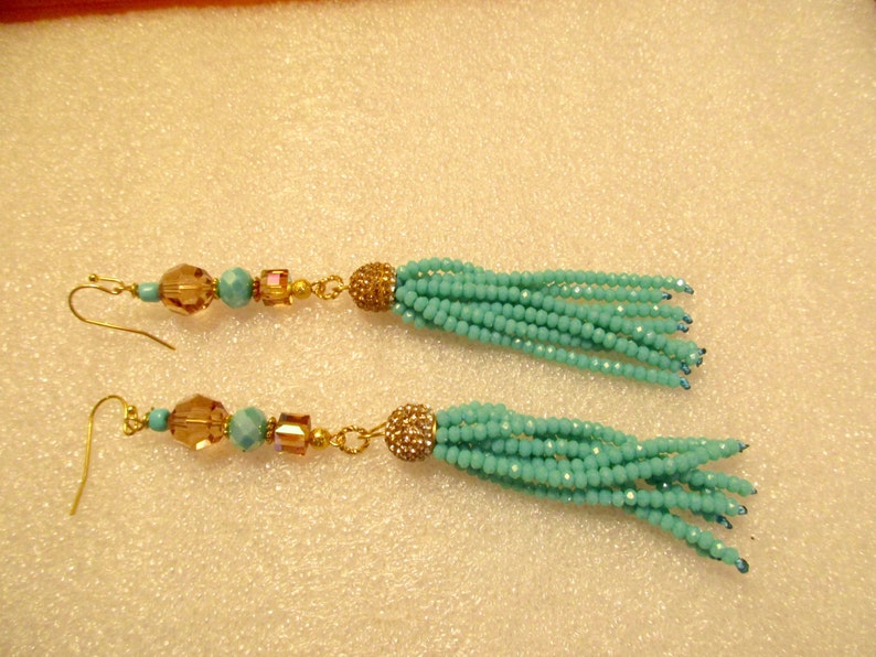 May include: A pair of long, beaded tassel earrings. The earrings feature gold-colored fish hooks, with a series of amber and turquoise beads leading to a cluster of turquoise beads. The tassels are made of small turquoise beads.