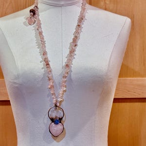 May include: A long, pink gemstone necklace with a heart-shaped pendant. The necklace is made of small, irregular pink stones. The pendant features a large pink heart, a blue stone, and a copper-colored metal ring.