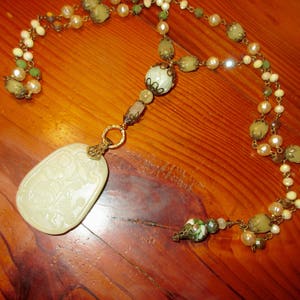 Puede incluir: Un collar con un colgante de jade blanco con dise&ntilde;o floral que cuelga de una cadena multicadena de cuentas verdes y blancas.