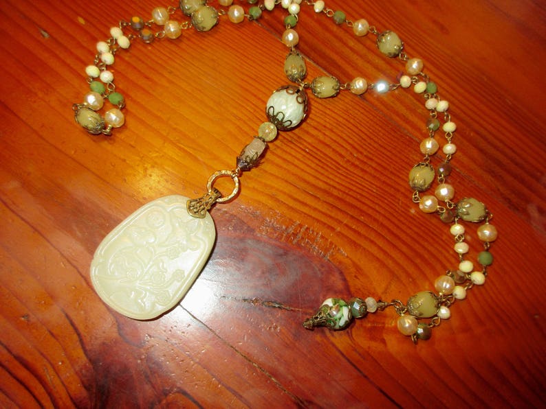 Puede incluir: Un collar con un colgante de jade blanco con dise&ntilde;o floral que cuelga de una cadena multicadena de cuentas verdes y blancas.
