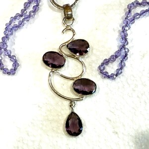May include: A silver necklace with a pendant featuring a swirling design with four amethyst stones. The necklace is made of small amethyst beads.