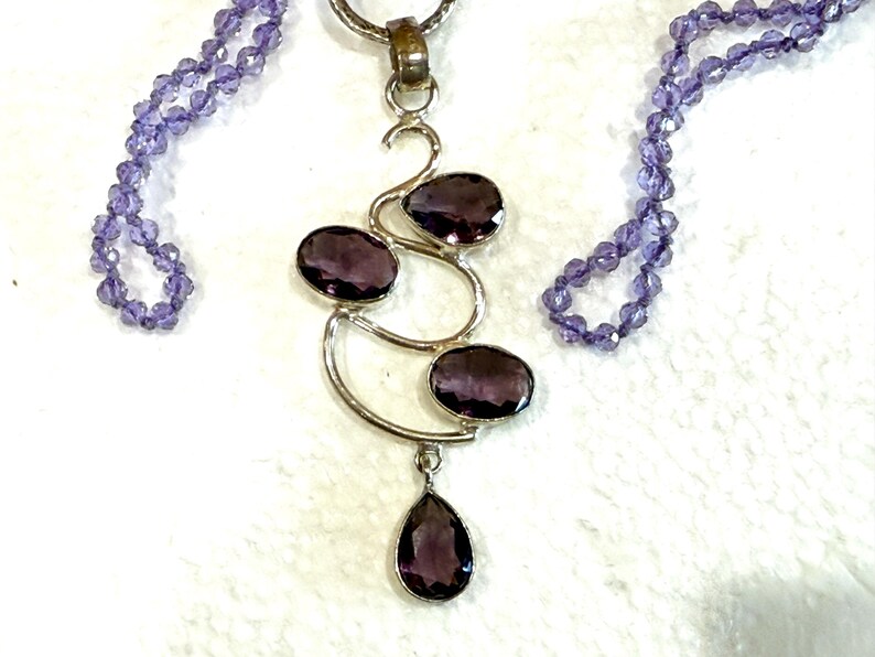 May include: A silver necklace with a pendant featuring a swirling design with four amethyst stones. The necklace is made of small amethyst beads.