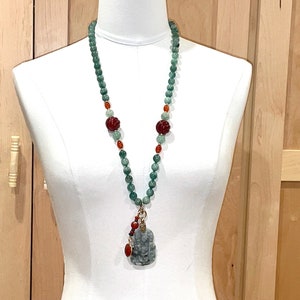 Puede incluir: Un collar largo de cuentas verdes y rojas con un colgante de jade con una figura tallada. El collar se muestra en un maniqu&iacute; blanco.