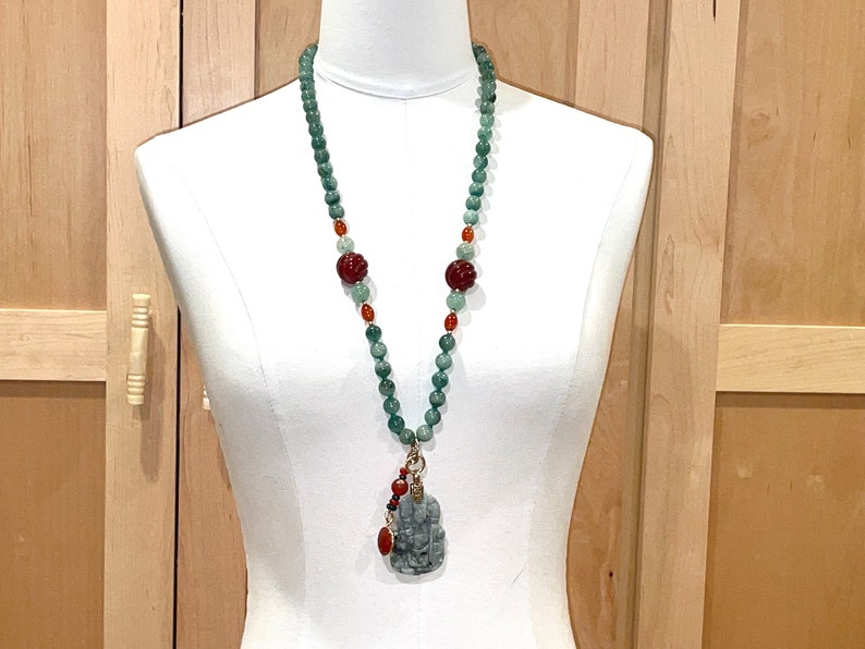 Puede incluir: Un collar largo de cuentas verdes y rojas con un colgante de jade con una figura tallada. El collar se muestra en un maniqu&iacute; blanco.