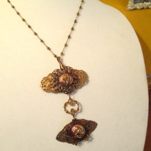 Może przedstawiać: Złoty naszyjnik z filigranowym wisiorem przedstawiającym r&oacute;żowo-fioletowy kwiat. Wisiorek jest przymocowany do mniejszego, podobnego wisiora z motywem motyla.