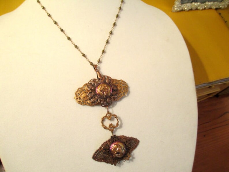 Może przedstawiać: Złoty naszyjnik z filigranowym wisiorem przedstawiającym r&oacute;żowo-fioletowy kwiat. Wisiorek jest przymocowany do mniejszego, podobnego wisiora z motywem motyla.