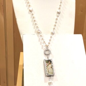 Pu&ograve; includere: Una collana di perle bianche con un ciondolo d'argento che presenta un'immagine incorniciata di una donna con un vestito bianco che tiene una rosa rosa.