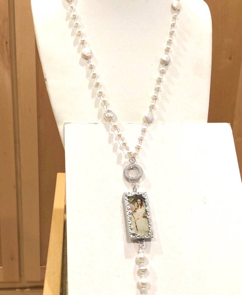 Pu&ograve; includere: Una collana di perle bianche con un ciondolo d'argento che presenta un'immagine incorniciata di una donna con un vestito bianco che tiene una rosa rosa.