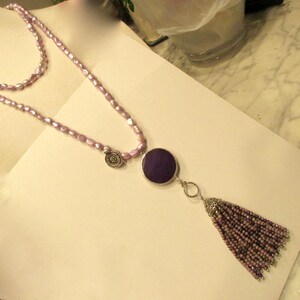 May include: A double-strand necklace made of light purple beads with a large, round, dark purple pendant and a tassel made of the same beads.