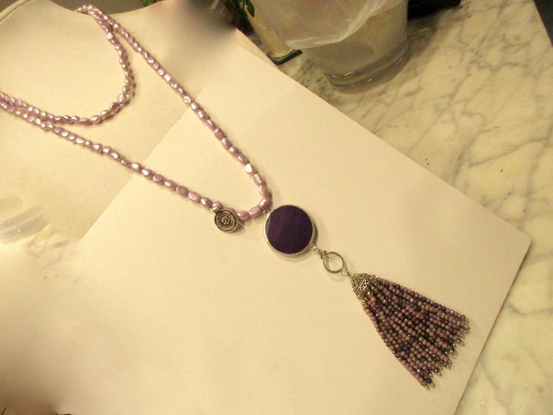 May include: A double-strand necklace made of light purple beads with a large, round, dark purple pendant and a tassel made of the same beads.