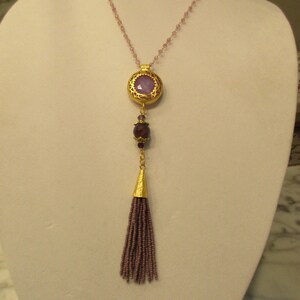 May include: A gold necklace with a purple stone pendant and a tassel. The necklace is made of small beads and has a delicate design.