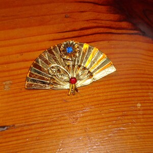 Puede incluir: Un broche de forma de abanico de color dorado con un dise&ntilde;o de pedrer&iacute;a azul y roja. El broche est&aacute; decorado con detalles intrincados y tiene una est&eacute;tica delicada y femenina.
