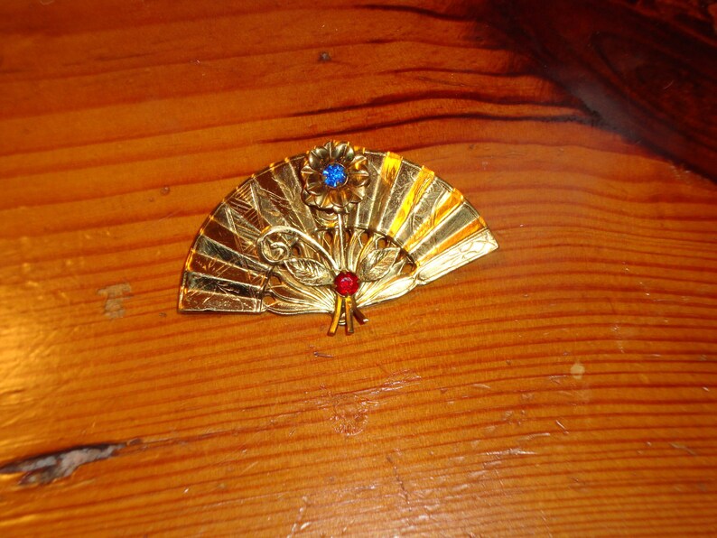 Puede incluir: Un broche de forma de abanico de color dorado con un dise&ntilde;o de pedrer&iacute;a azul y roja. El broche est&aacute; decorado con detalles intrincados y tiene una est&eacute;tica delicada y femenina.