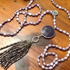 May include: A long lavender pearl necklace with a large, faceted, purple gemstone pendant and a tassel of small, gray beads.
