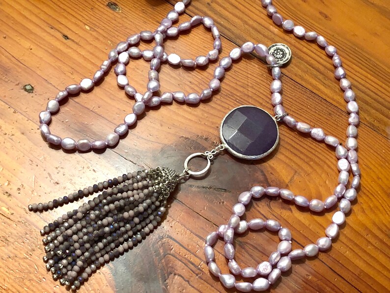 May include: A long lavender pearl necklace with a large, faceted, purple gemstone pendant and a tassel of small, gray beads.