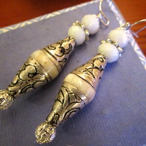 May include: A pair of silver dangle earrings with white beads and intricate silver designs. The earrings have a teardrop shape and are accented with small silver beads.