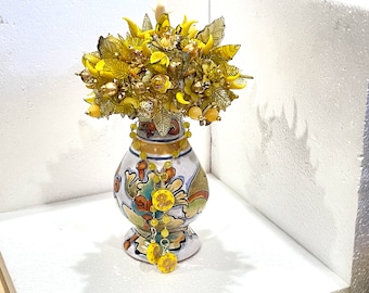 May include: A decorative arrangement of yellow glass flowers and leaves in a ceramic vase with a painted floral design. A matching necklace and earrings with yellow beads and circular pendants hang from the vase.