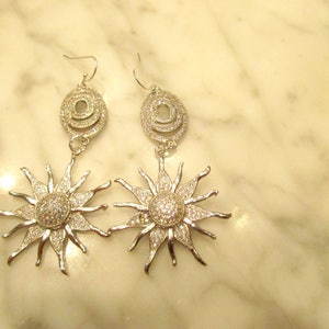 May include: Silver tone dangle earrings with a sunburst design and a circle of clear stones. The earrings are on a white marble surface.