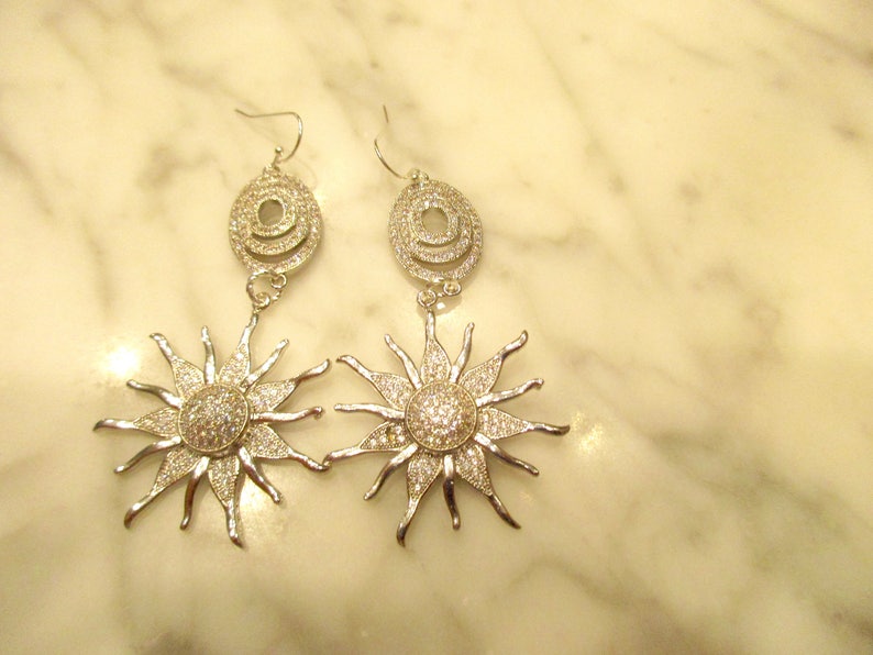 May include: Silver tone dangle earrings with a sunburst design and a circle of clear stones. The earrings are on a white marble surface.