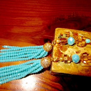 May include: A pair of turquoise beaded tassel earrings with gold-colored accents. The earrings feature a cluster of small, round beads in a vibrant turquoise color, topped with a gold-colored, glittery ball. The earrings are on a wooden surface.