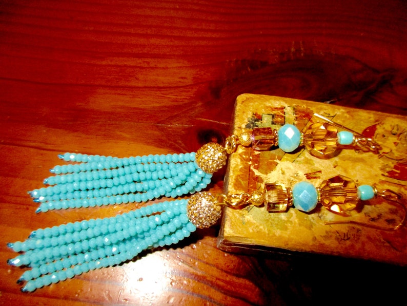 May include: A pair of turquoise beaded tassel earrings with gold-colored accents. The earrings feature a cluster of small, round beads in a vibrant turquoise color, topped with a gold-colored, glittery ball. The earrings are on a wooden surface.