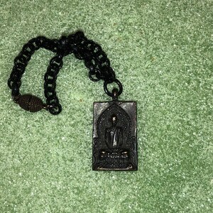 May include: A black chain necklace with a rectangular pendant featuring a black and silver image of a seated Buddha. The pendant has Thai text on the bottom.