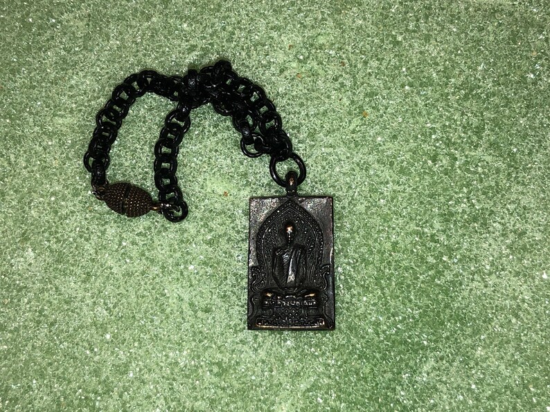 May include: A black chain necklace with a rectangular pendant featuring a black and silver image of a seated Buddha. The pendant has Thai text on the bottom.