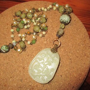 Puede incluir: Un collar con un colgante de jade verde y blanco con un dise&ntilde;o floral tallado. El collar est&aacute; hecho con cuentas verdes y blancas y una cadena dorada.