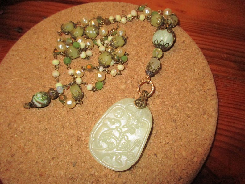 Puede incluir: Un collar con un colgante de jade verde y blanco con un dise&ntilde;o floral tallado. El collar est&aacute; hecho con cuentas verdes y blancas y una cadena dorada.