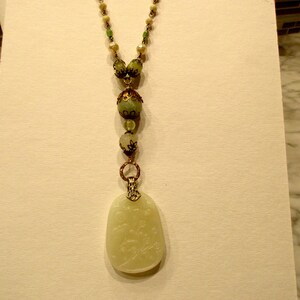 Puede incluir: Un collar largo de cuentas verdes y blancas con un colgante de jade tallado. El colgante tiene forma ovalada y presenta un dise&ntilde;o floral.