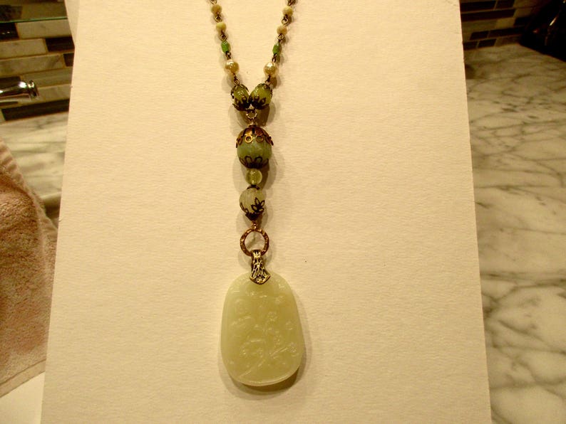 Puede incluir: Un collar largo de cuentas verdes y blancas con un colgante de jade tallado. El colgante tiene forma ovalada y presenta un dise&ntilde;o floral.