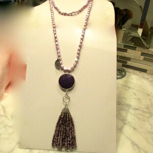 May include: A long, layered necklace with a purple tassel and a round, purple pendant. The necklace is made of small, light purple beads.