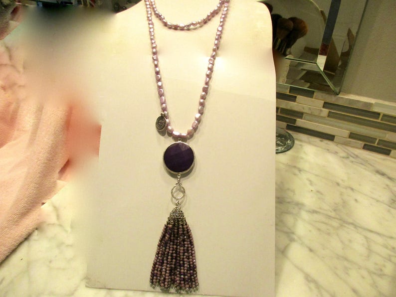 May include: A long, layered necklace with a purple tassel and a round, purple pendant. The necklace is made of small, light purple beads.