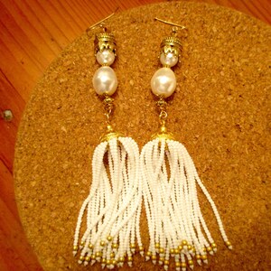 May include: A pair of white beaded tassel earrings with gold accents. The earrings feature a large white pearl and a gold cap at the top.