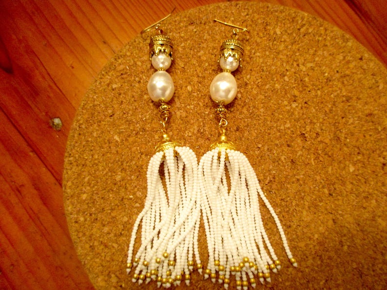 May include: A pair of white beaded tassel earrings with gold accents. The earrings feature a large white pearl and a gold cap at the top.