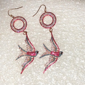 Puede incluir: Un par de pendientes colgantes con un dise&ntilde;o de p&aacute;jaro rosa y azul. Los pendientes tienen un aro redondo engastado con cristales y un colgante de p&aacute;jaro. Los p&aacute;jaros est&aacute;n adornados con cristales rosas y azules.